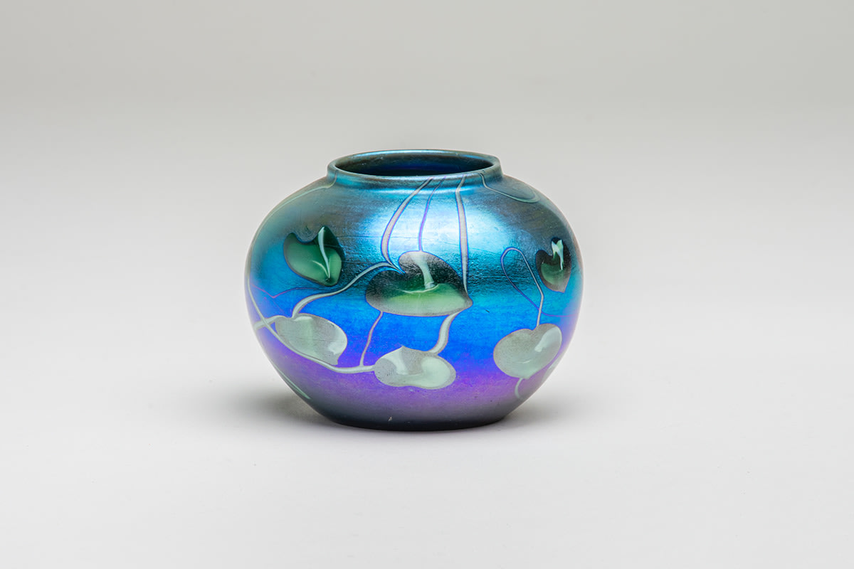 a round blue iridescent cabinet vase by Tiffany Studios, the favrile glass vase decorated with a motif of pointed green leaves with thin swirling and trailing vines