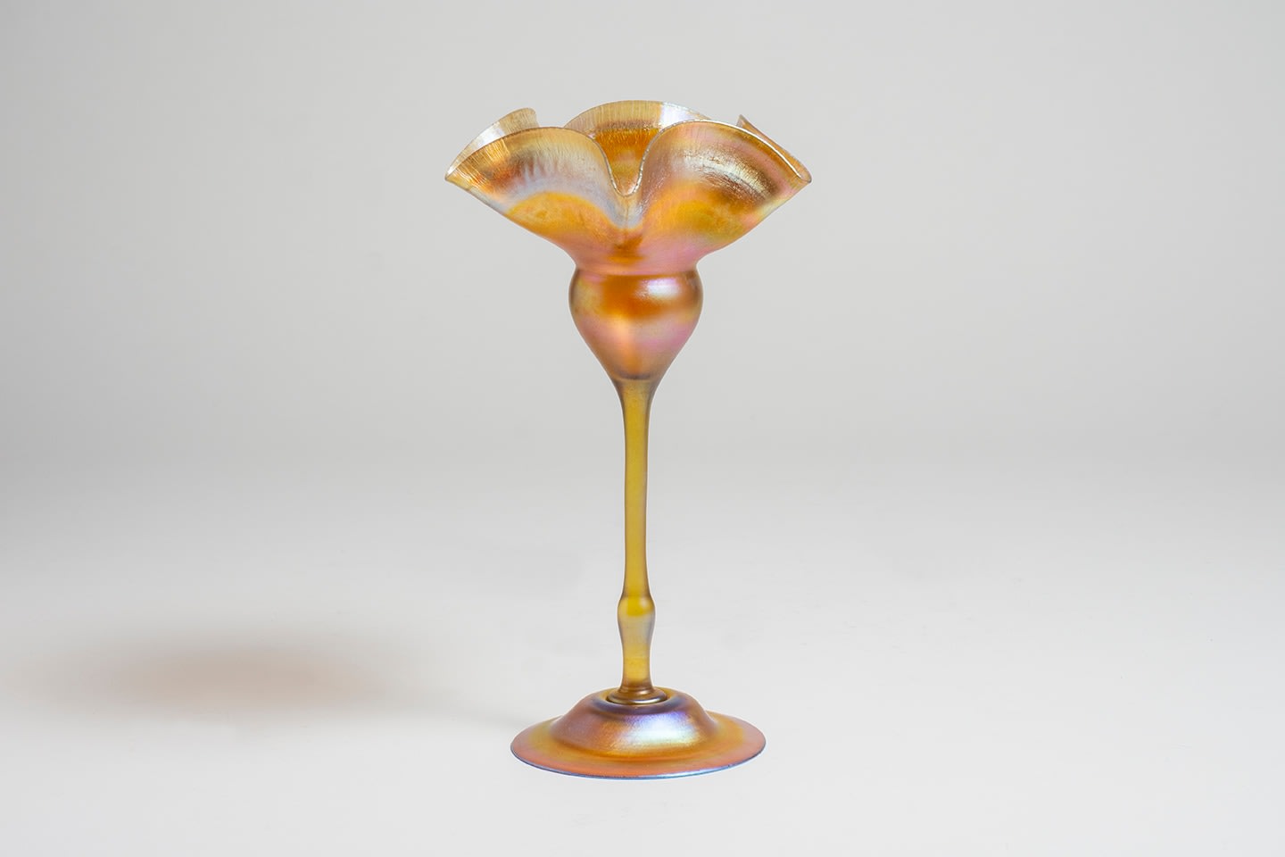 a gold iridescent tiffany glass flower form vase, a domed foot supporting thin stem which rises to support a rounded cup with pinched heavily ruffled rim