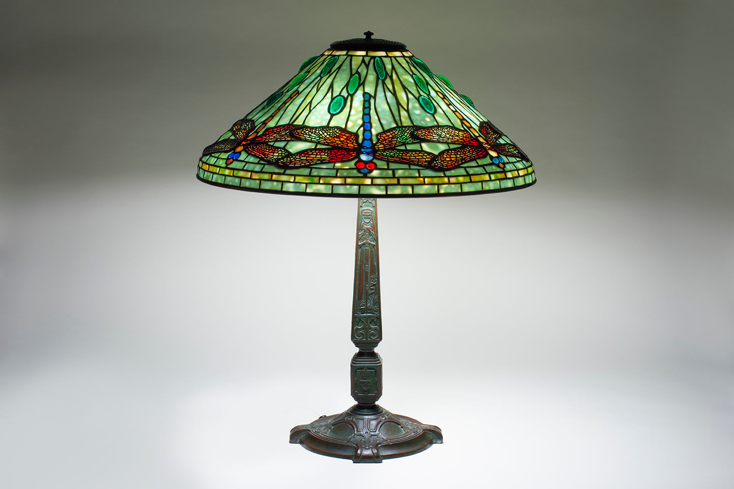 a leaded glass original tiffany lamp with dragonfly shade, depicting a series of downward facing dragonflies in a variety of glass colors ranging from green, blue and red, with rounded glass gems for the eyes, against a background of mottled glass in a shade of pale blue-green