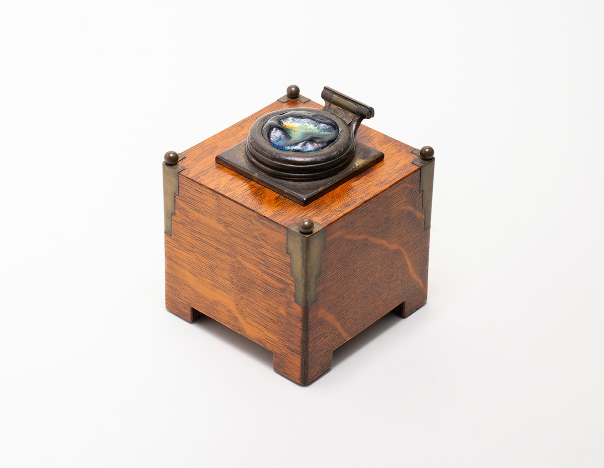 a squared oak inkwell in quarter sawn oak with highly figured grain, the top with small circular inset inkwell with hinged metal lid inset with a pressed Tiffany glass &quot;turtle back&quot; tile in iridescent blue