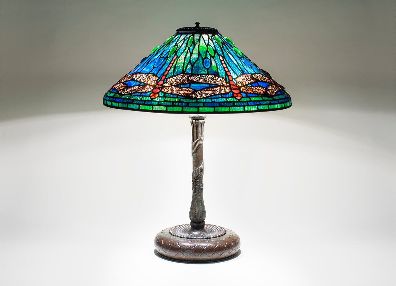 a bronze and leaded glass tiffany lamp dating from 1906, depicting pink and red dragonflies with pierced filigree wings floating above a background of translucent rippled glass in shades of blue representing water