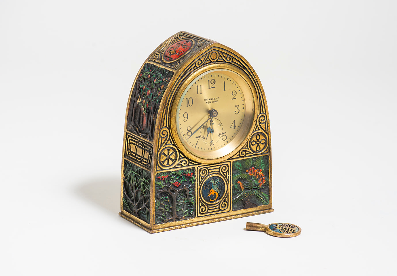 a desk clock with rectangular base and pointed top, the round clock face marked TIFFANY &amp; CO, from the &quot;Bookmark&quot; Desk set produced by Tiffany Studios around 1910, in gilt bronze with areas of sculptural depiction in relief of stylized monograms and natural motifs like palm trees, pine needs meant to represent Renaissance printer's marks
