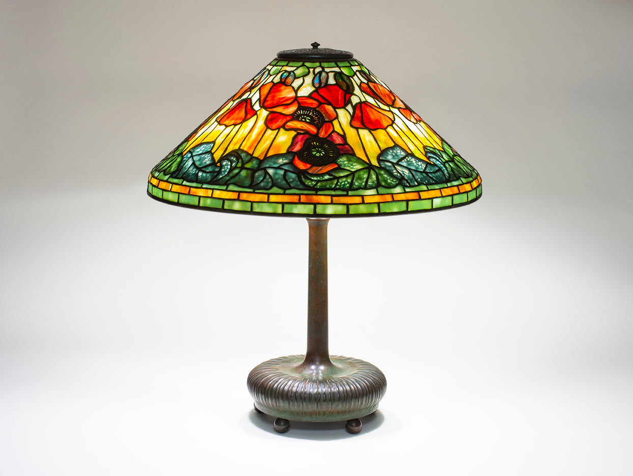 a leaded glass Tiffany Lamp depicting red poppies with pierced metal filigree mounted on top of the glass representing the centers of the flowers, against a background of variegated yellow glass, with poppy buds in blue-red glass near the top of the shade, the lower edge of the shade with green leaves, the vines formed by pierced metalwork mounted to the interior of the shade.