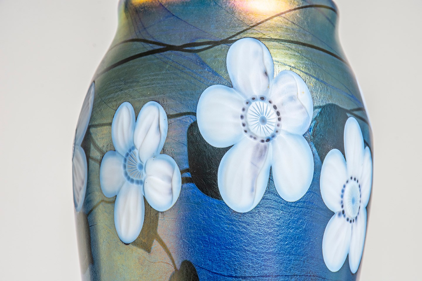 a detail of a blue tiffany favrile glass vase, the floral decoration in white with the center and petals carved with natural details, against a deep blue background glass with silvery surface iridescence