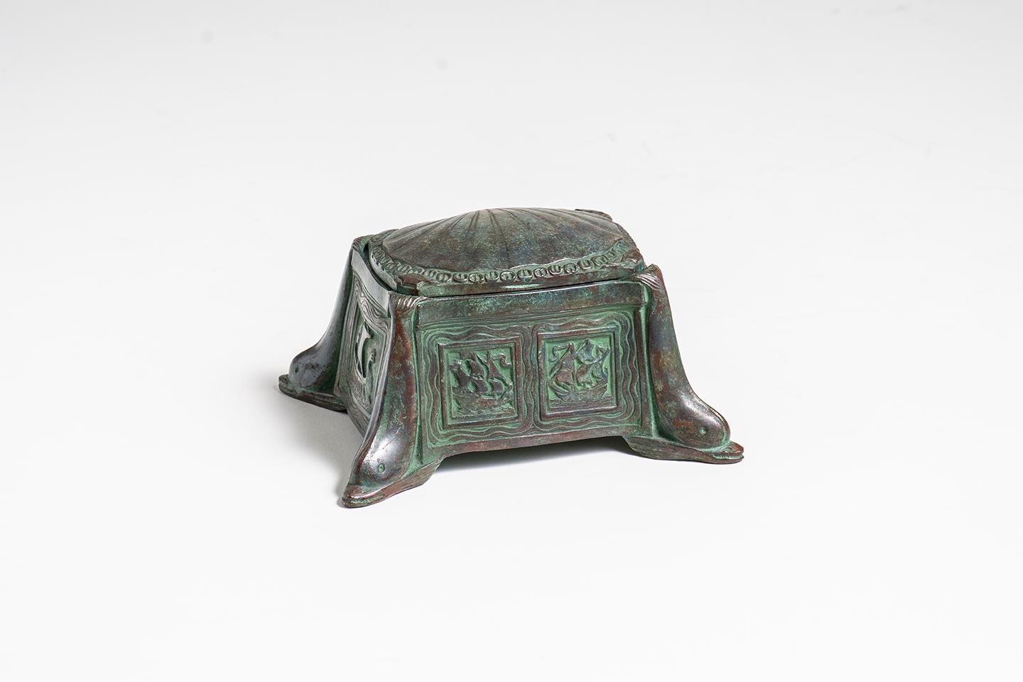 a rectangular bronze inkwell with soft brow-green patina from the Nautical Desk Set by Tiffany Studios which featured &quot;varied motifs of the sea,&quot; this inkwell with low relief depictions of sailing ships on each of the four sides, the top formed by a hinged scallop shell, each of the four corners decorated with a diving dolphin