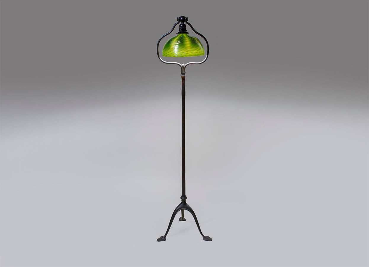 a tiffany studios floor lamp featuring green blown glass shade with overall iridescent wave design in silvery blue, on a three-footed floor base with thin stem and harp top which allows the shade to be swiveled.