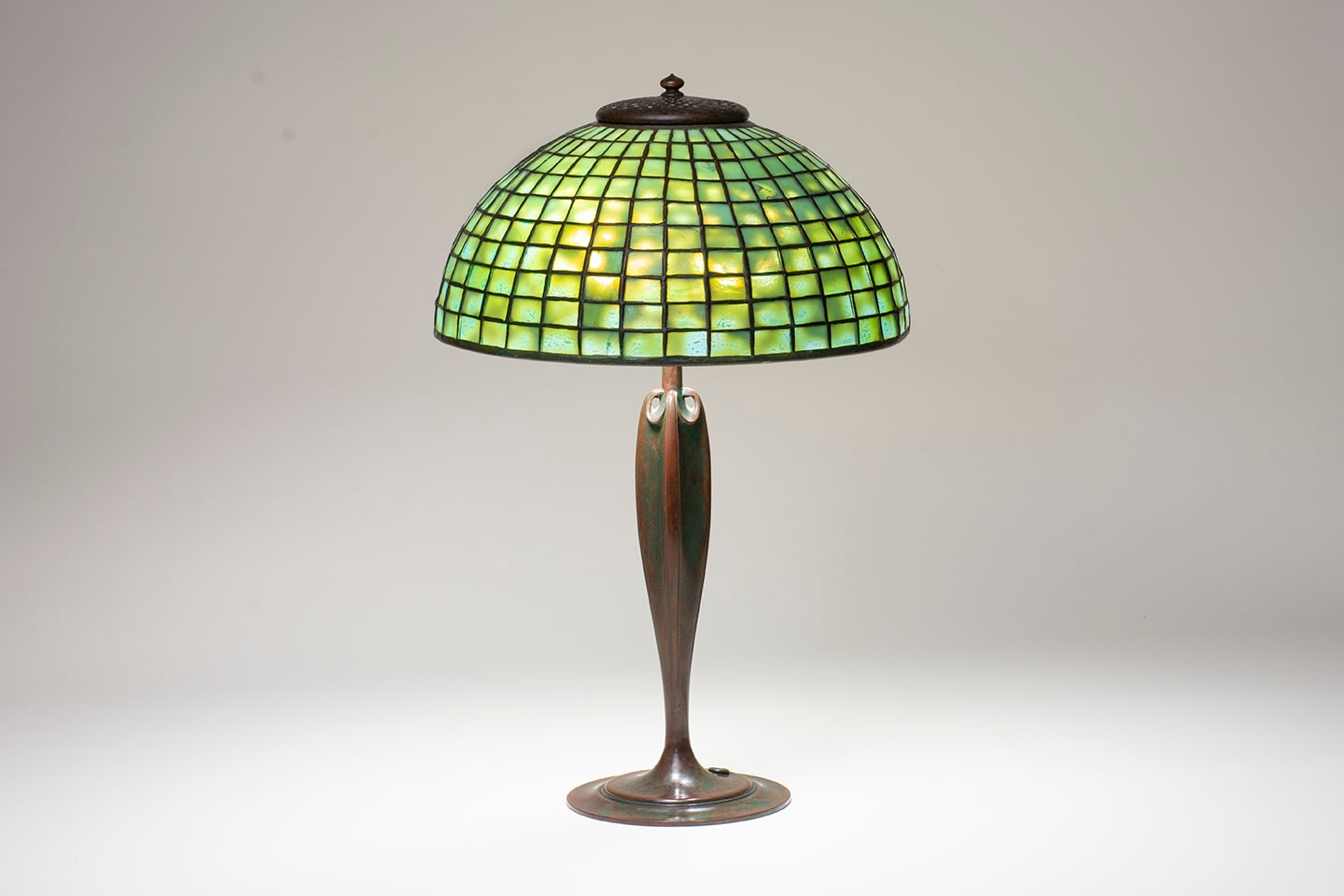 an authentic Tiffany Lamp dating from the early 20th century, pictured with the shade illuminated, with an elegant Art Nouveau style bronze base in rich brown patina, supporting a dome-shaped leaded glass shade formed by square shaped pieces of heavily mottled Tiffany Glass