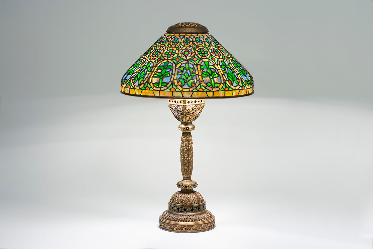 an original tiffany lamp from the venetian desk set, the complex gilt bronze base matching the desk set's intricate openwork pattern of scrollwork and motif of ermines, with inset green rounded glass jewels, paired with a conical shaded of leaded tiffany glass depicting alternating medallions of palmettes and fleur de lys with a geometric motif in blue, red, and green glass