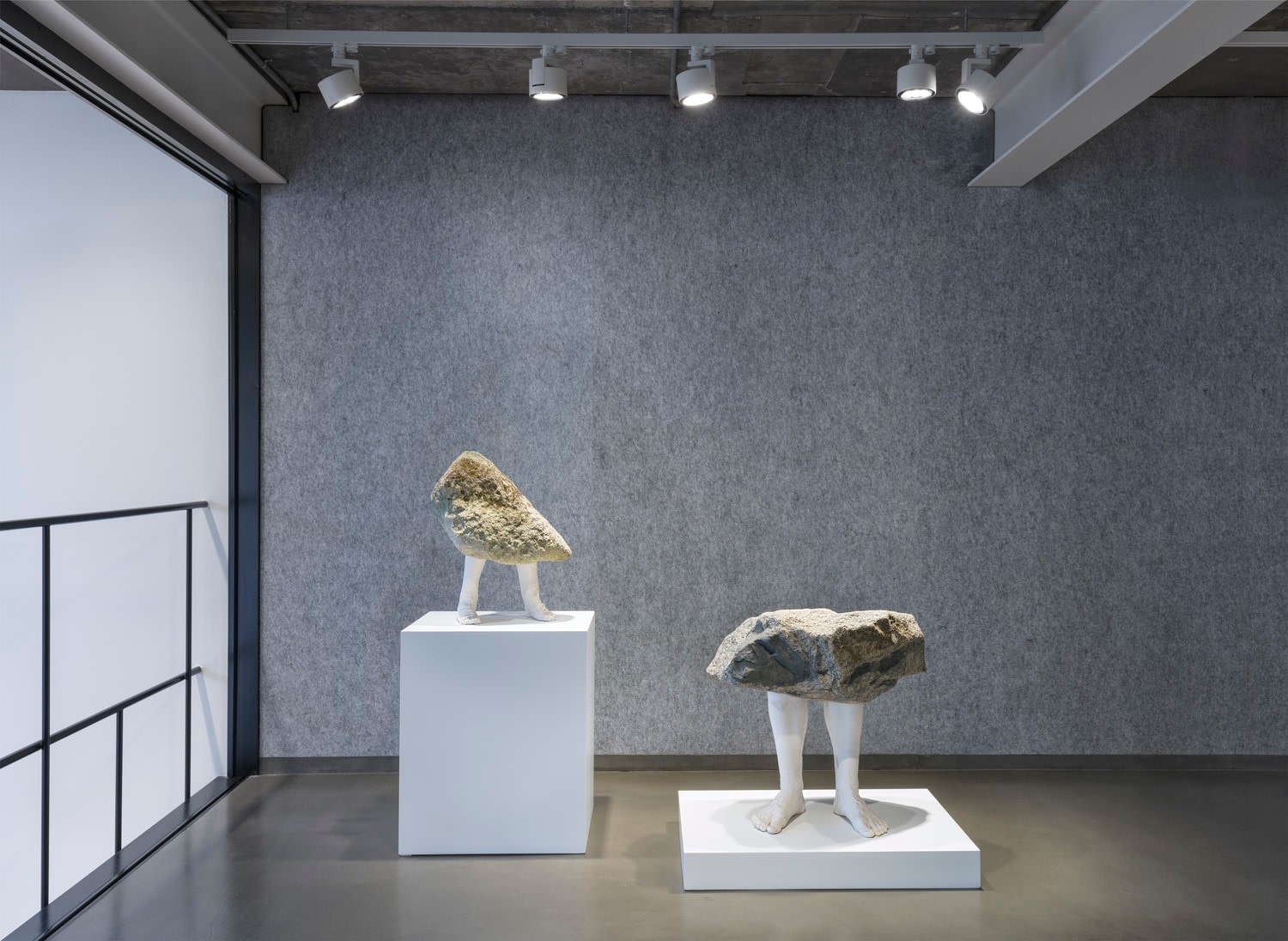 Two sculptures of rocks on legs each standing on platforms one higher than the other in front of a grey gallery wall