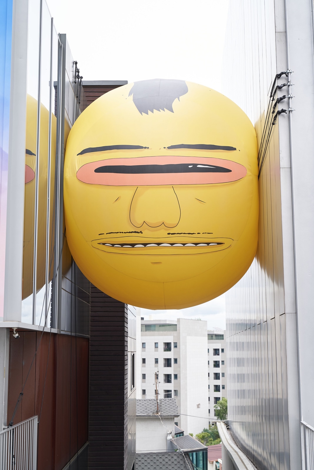 Exterior view of the exhibition OSGEMEOS: You Are My Guest in Seoul. Large yellow balloon with a face squeezed between two buildings
