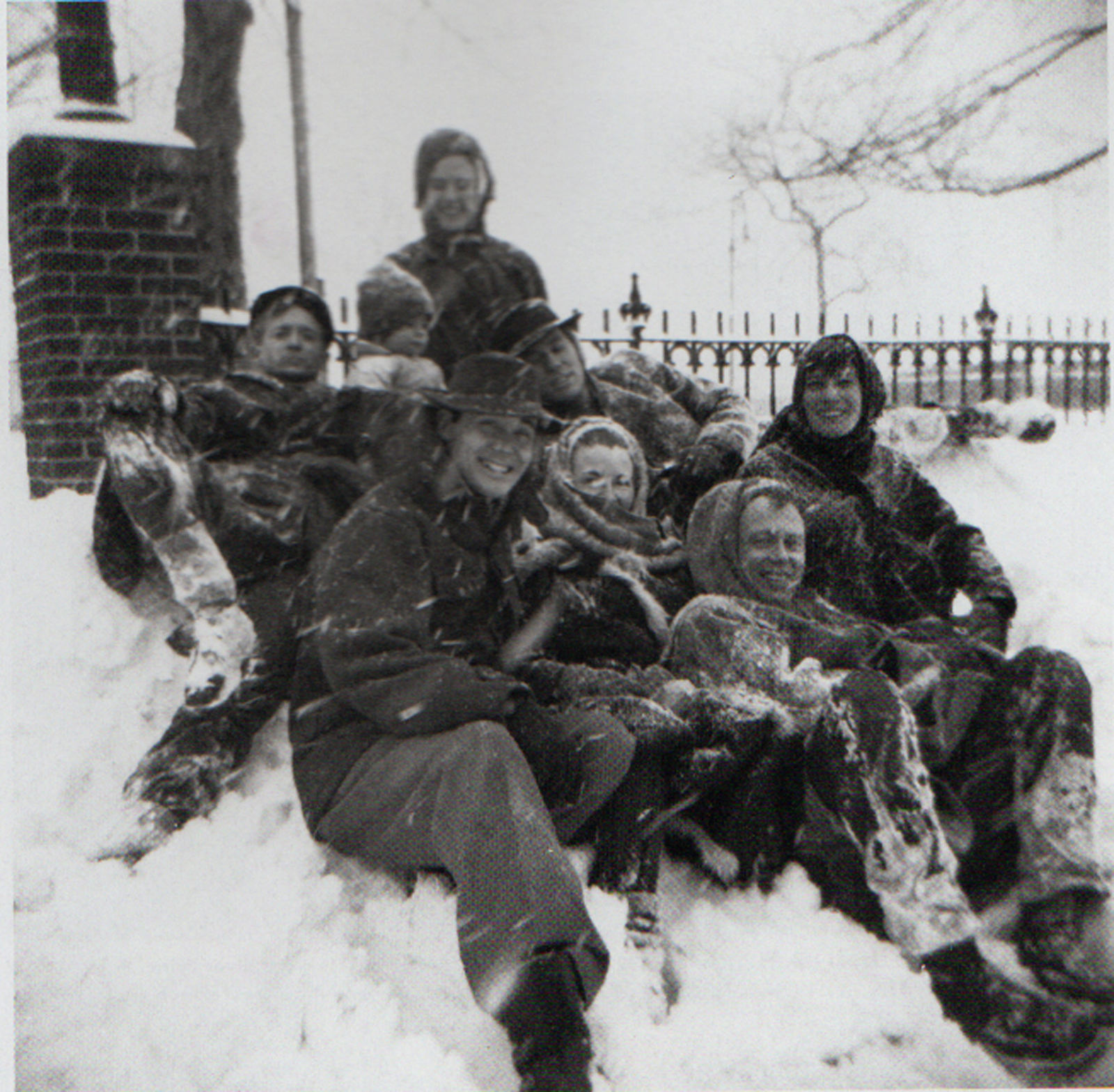A black and white photograph of Jack Youngerman, Duncan Youngerman, Delphine Seyrig, Jerry Matthews, Dolores Matthews, Ellsworth Kelly, Lenore Tawney, and Robert Clark sitting in the snow in Jeannette Park, New York on February 15, 1958