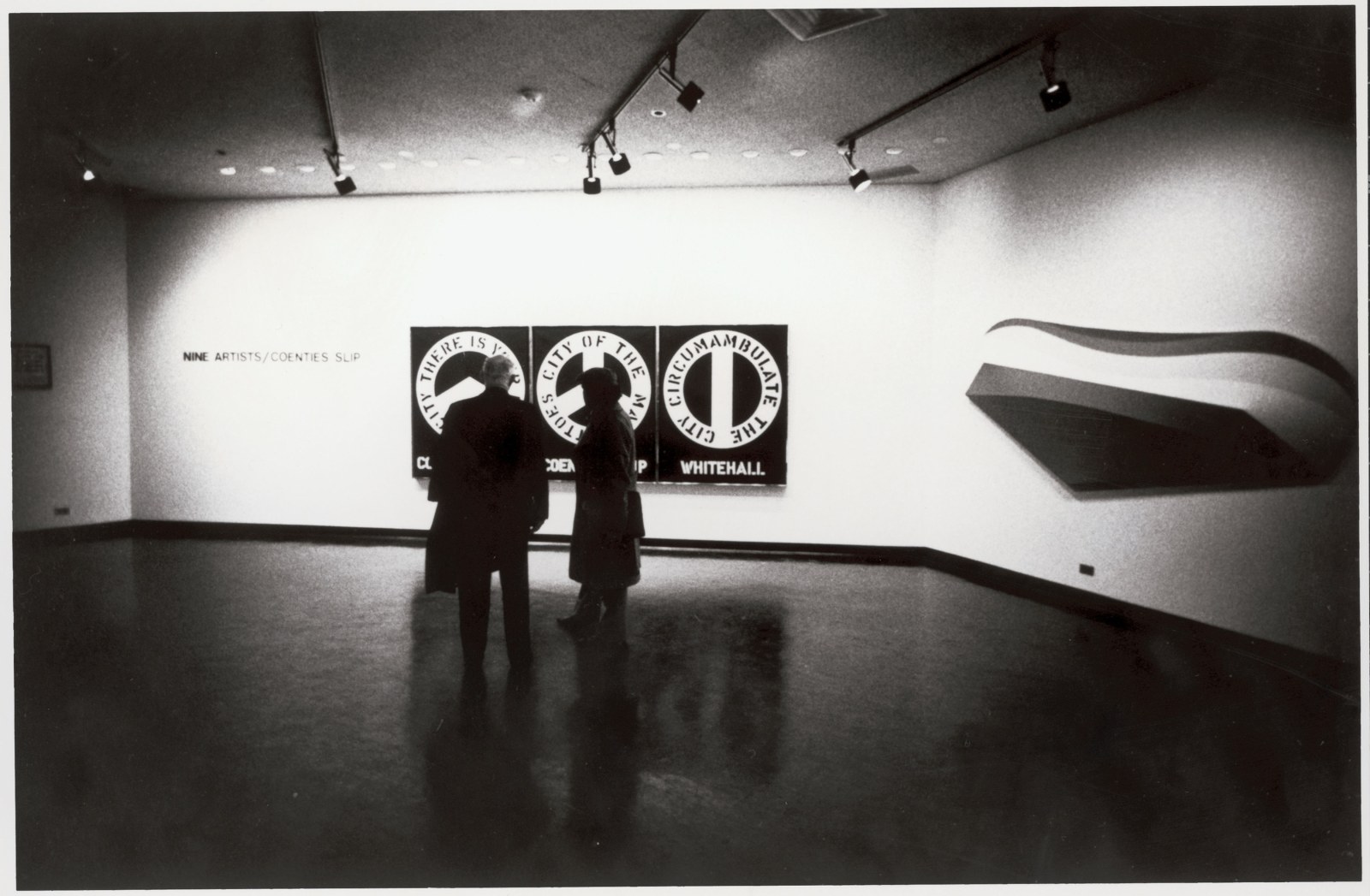 A black and white photo of a man and woman in front of Indiana’s painting The Melville Triptych (1961) in the exhibition Nine Artists: Coenties Slip at the Whitney Museum of American Art Downtown, New York, January 10–February 14, 1974. Also visible is Charles Hinman’s Lift (1965)