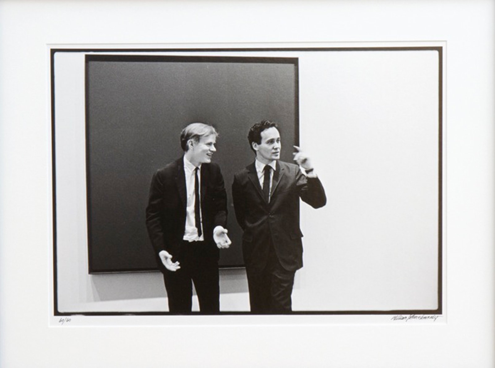 Black and white photograph of Andy Warhol and Indiana in front of an unidentified canvas by Ad Reinhardt at the opening of the exhibition Americans 1963 at the Museum of Modern Art, New York, May 21, 1963