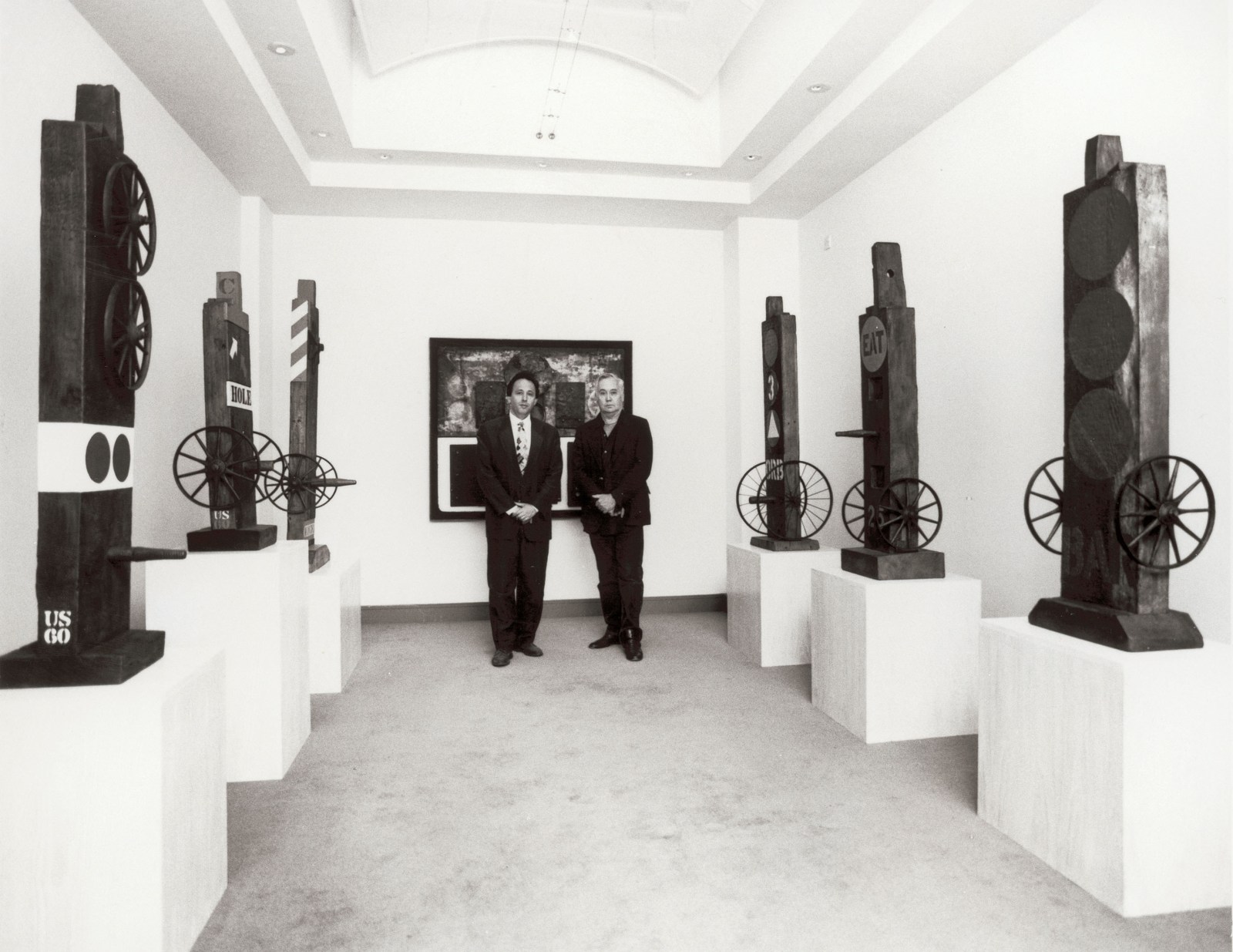 Black and white photograph of Simon Salama-Caro and Robert Indiana in front of the wall construction Wall of China at the Salama-Caro Gallery, London, during the exhibition Robert Indiana: Early Sculpture, 1960–1962, in September 1991. Three sculptures are displayed to the left and right of them.