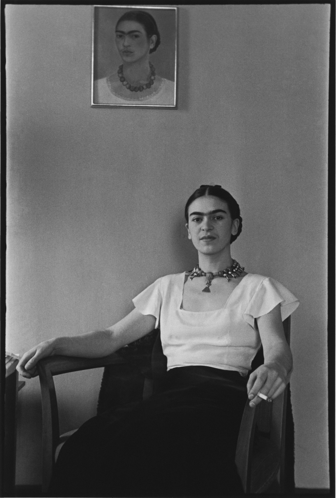 Frida Kalho sits in a chair and looks at the camera while holding a cigarette and sitting beneath one of her self-portraits.