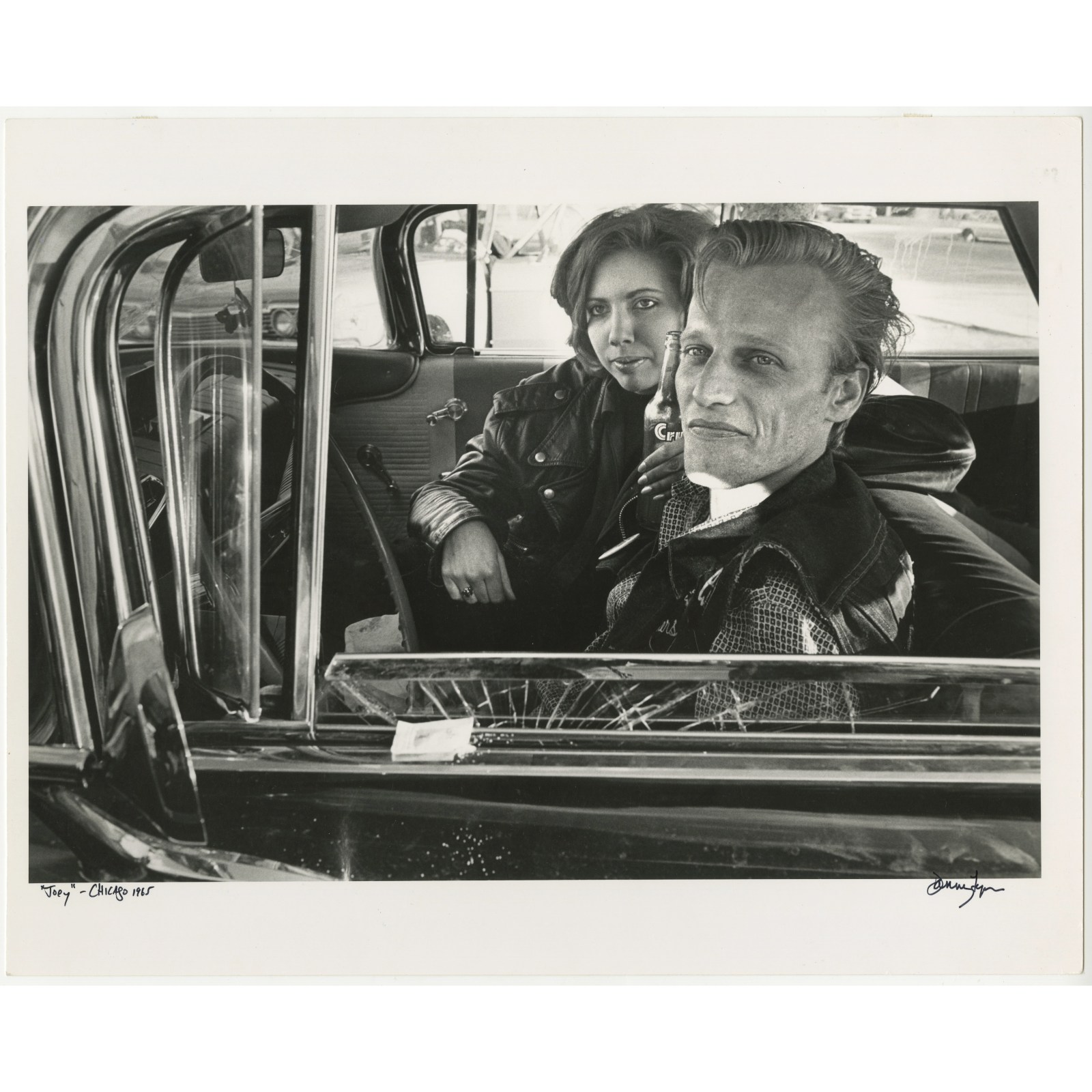 A couple sit in an old car while wearing leather jackets and looking towards the photographer in this black and white photograph.