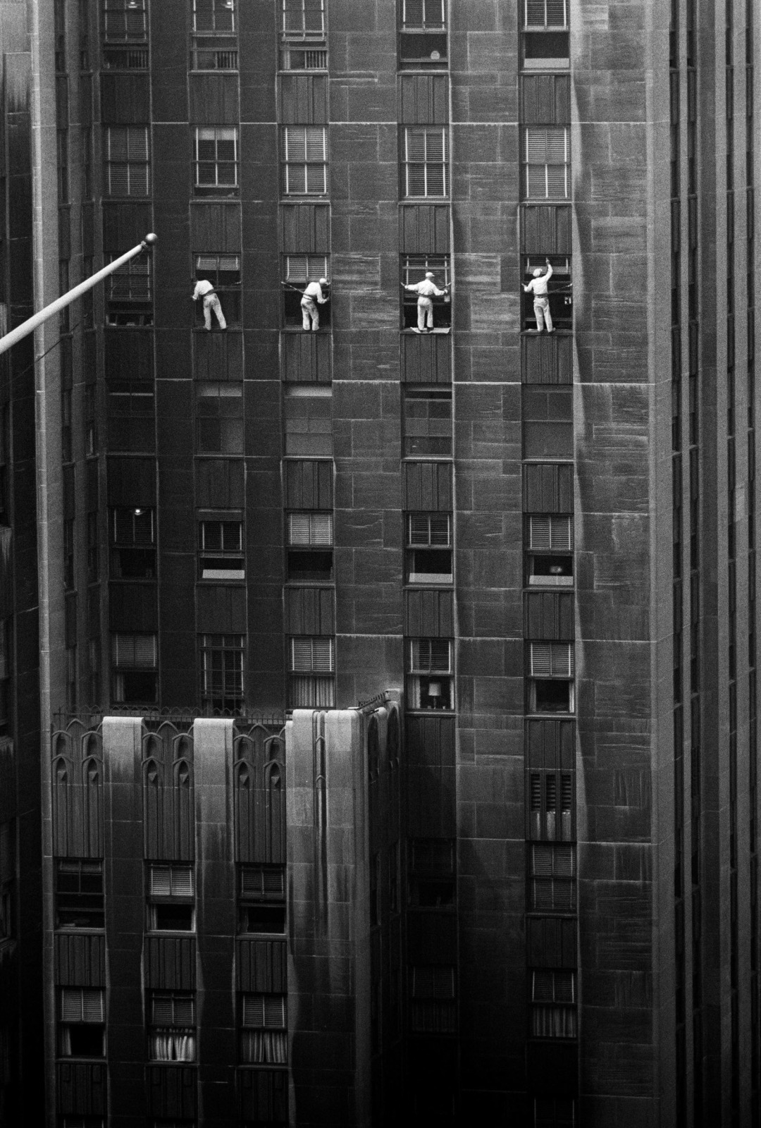 Four window-washers wearing white are photographed in black and white on a skyscraper in motion as they clean the windows.