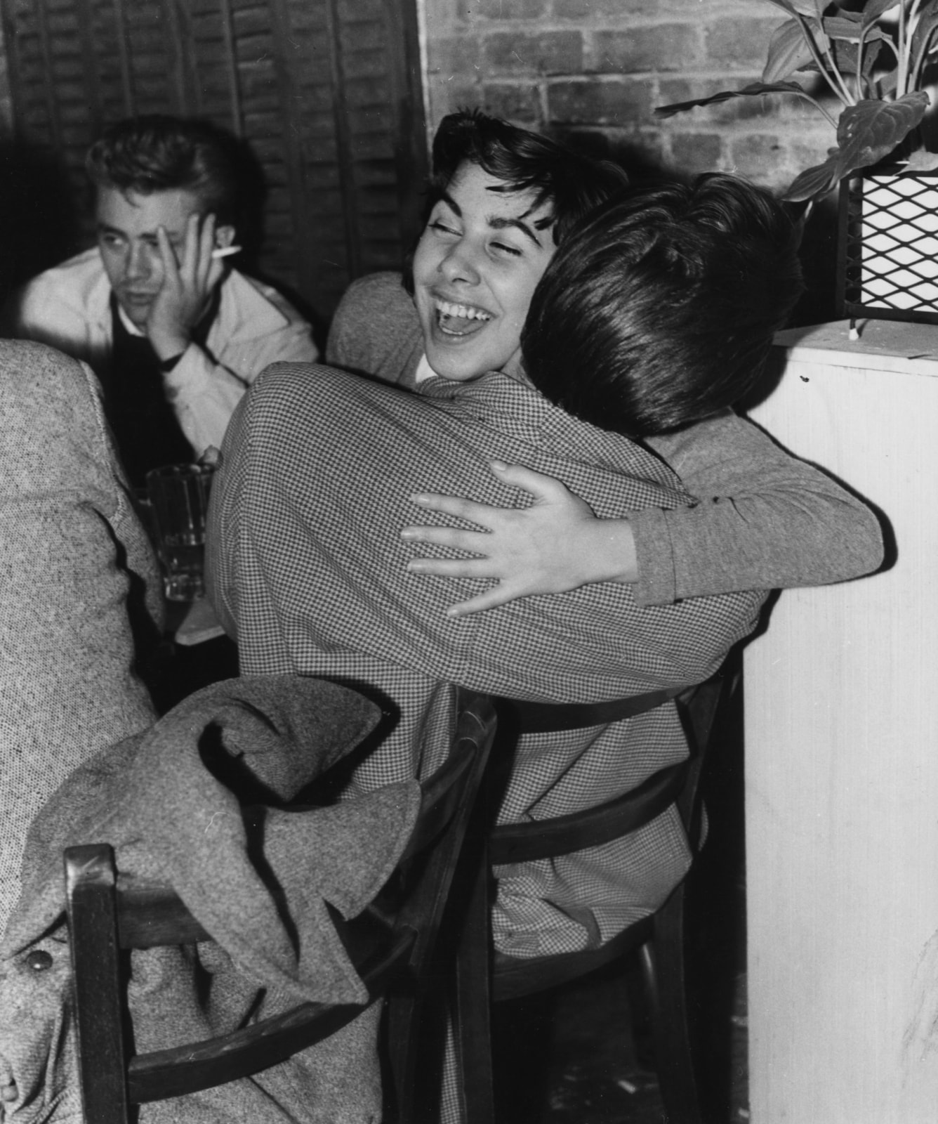 A young woman smiles as she hugs a man while actor James Dean smokes and looks away, bored, in this black and white photo.