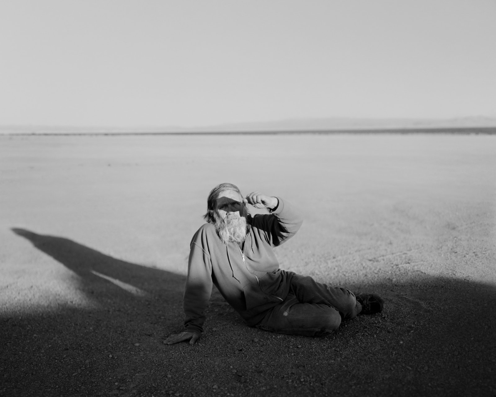 A man is photographed in black and white in a desolate desert-like landscape, as he sits on the ground and shields his eyes from the sun.