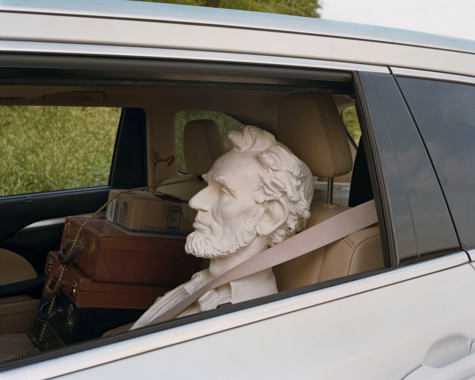 A sculpture of Abe Lincoln strapped into the back seat of a car with small briefcases stacked in the car next to him