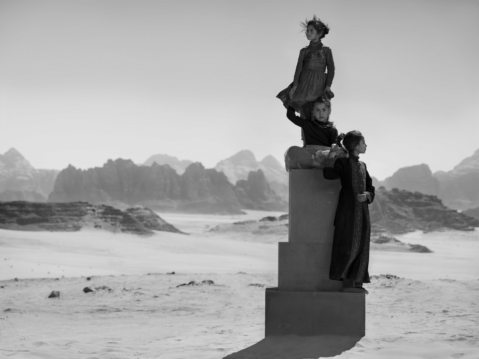 Three young girls are photographed in the desert while wearing traditional dress and standing at various levels of a pediment.