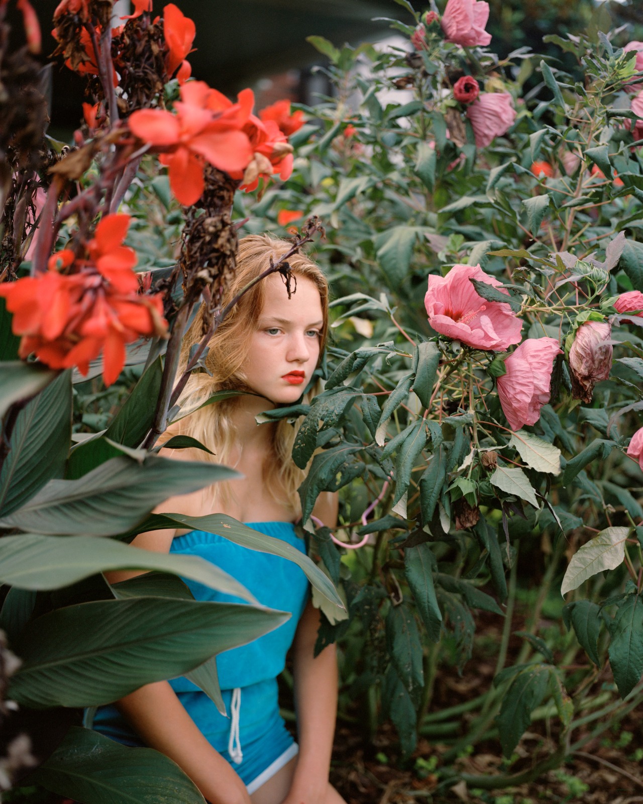 A young girl in a teal romper wears bring reddish-orange lipstick and sits amongst tall colorful flowers while staring into the near-distance.