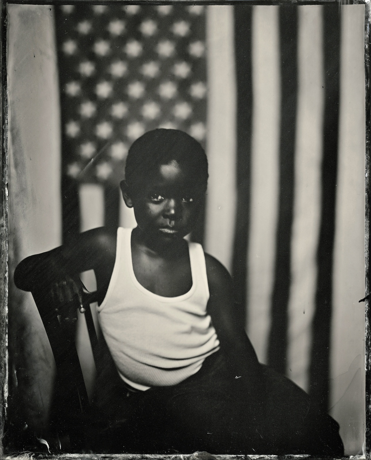 A Black child sits in front of the American flag and looks into the camera in this black and white photograph
