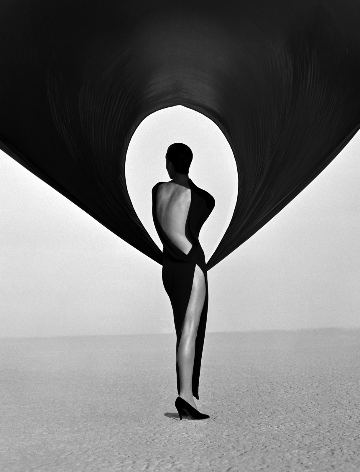 A black and white photo of a figure in a black dress in the desert with black fabric suspended overhead