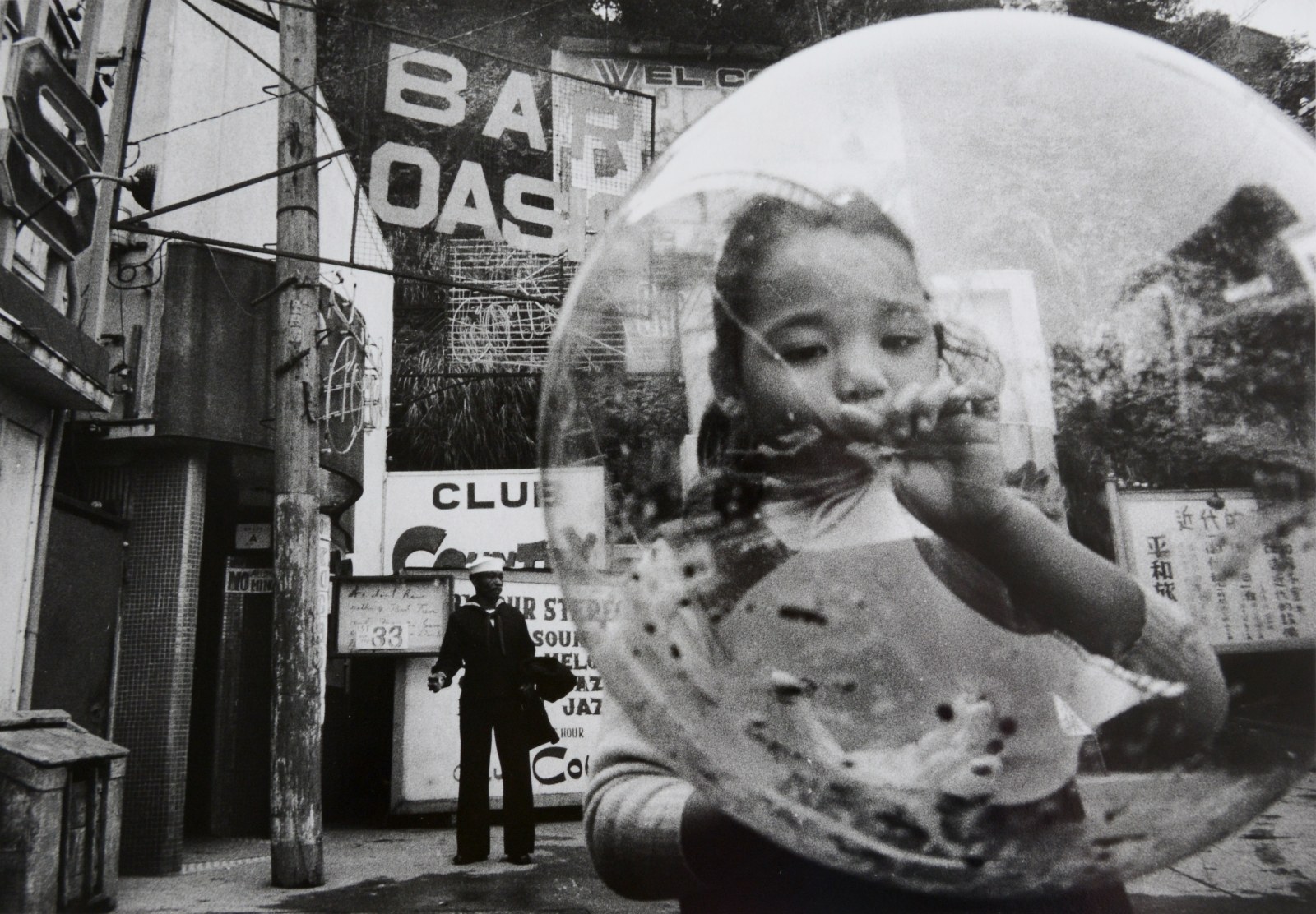 A child blows up a large, clear balloon and a sailor stands in the distance against a wall in a black and white photo