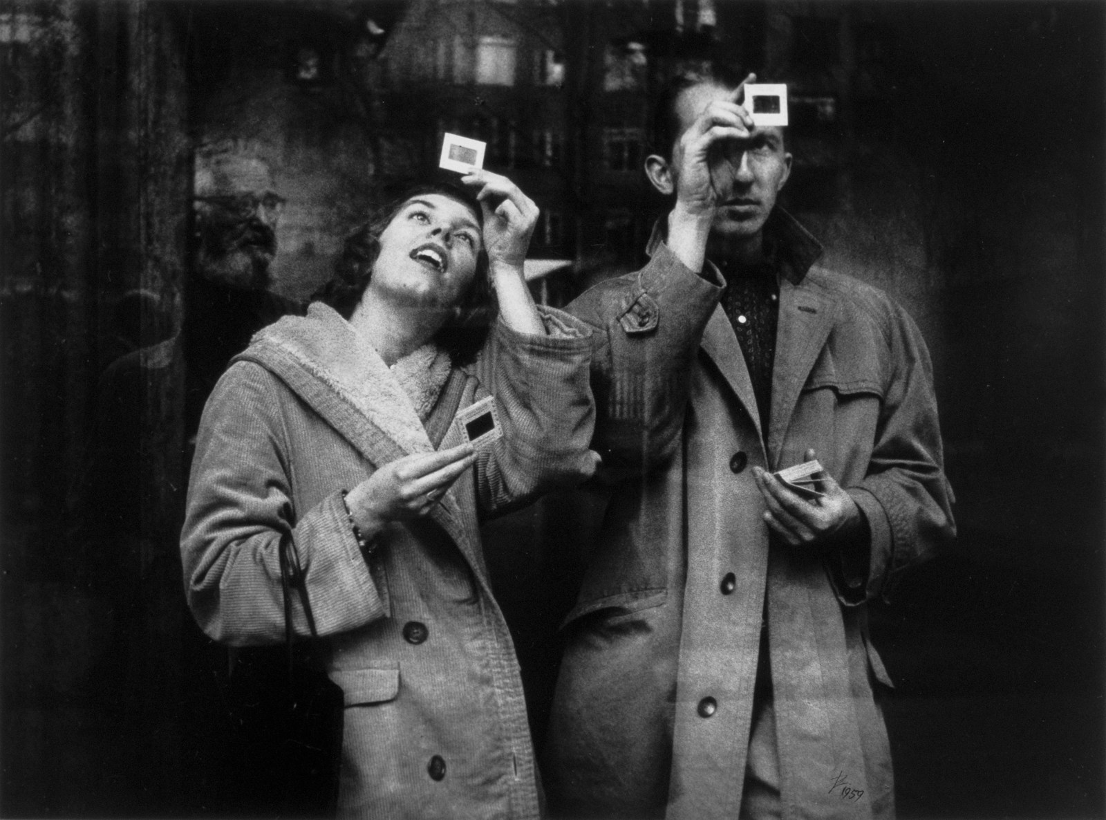 A man and a woman photographed in black and white hold up slides and look through them to see the image