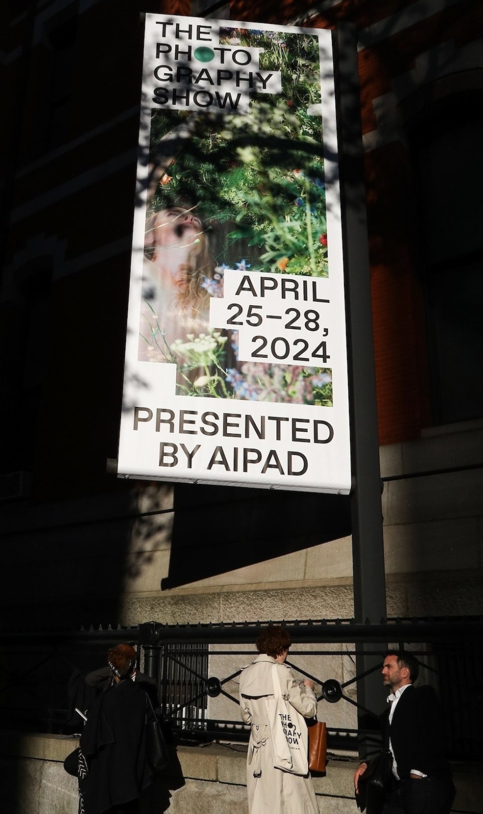 The show banner is lit by the sun through dappling trees while people stand outside The Photography Show 2024