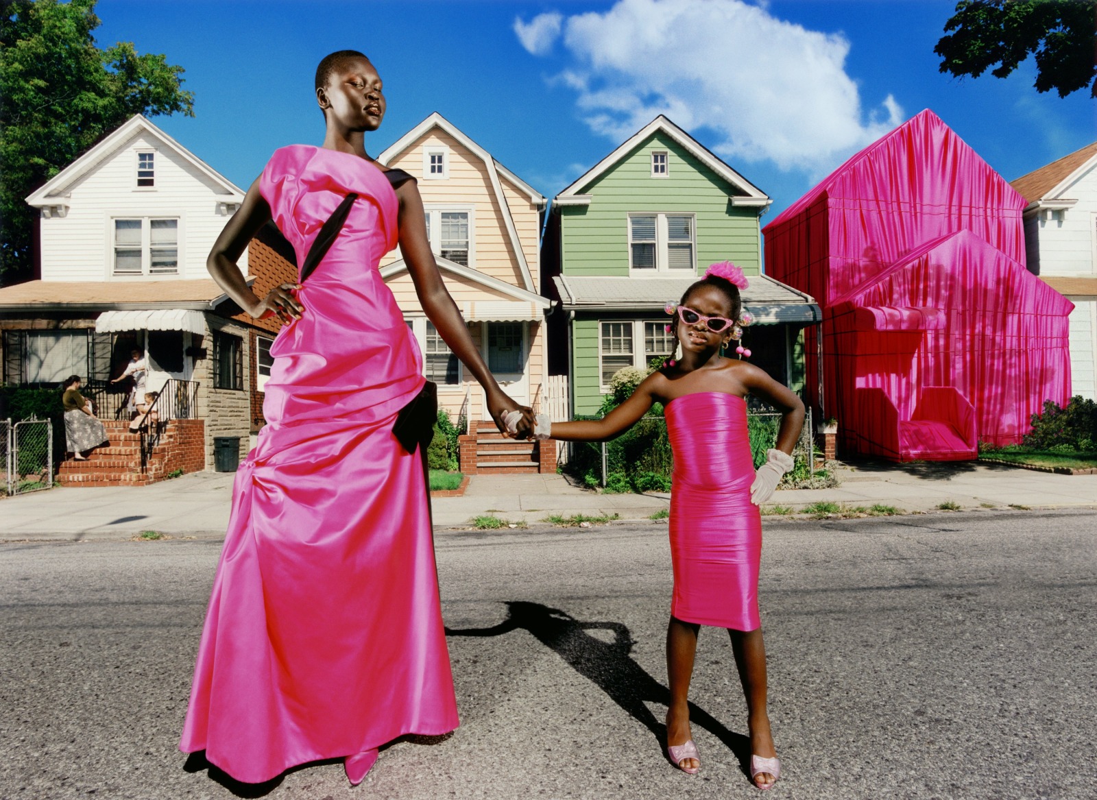 A tall Black female model stands in a hot pink formal dress on a street with houses, while holding the hand of a little girl's hand. The girl is also wearing hot pink and one of the houses in the background is draped in hot pink fabric.