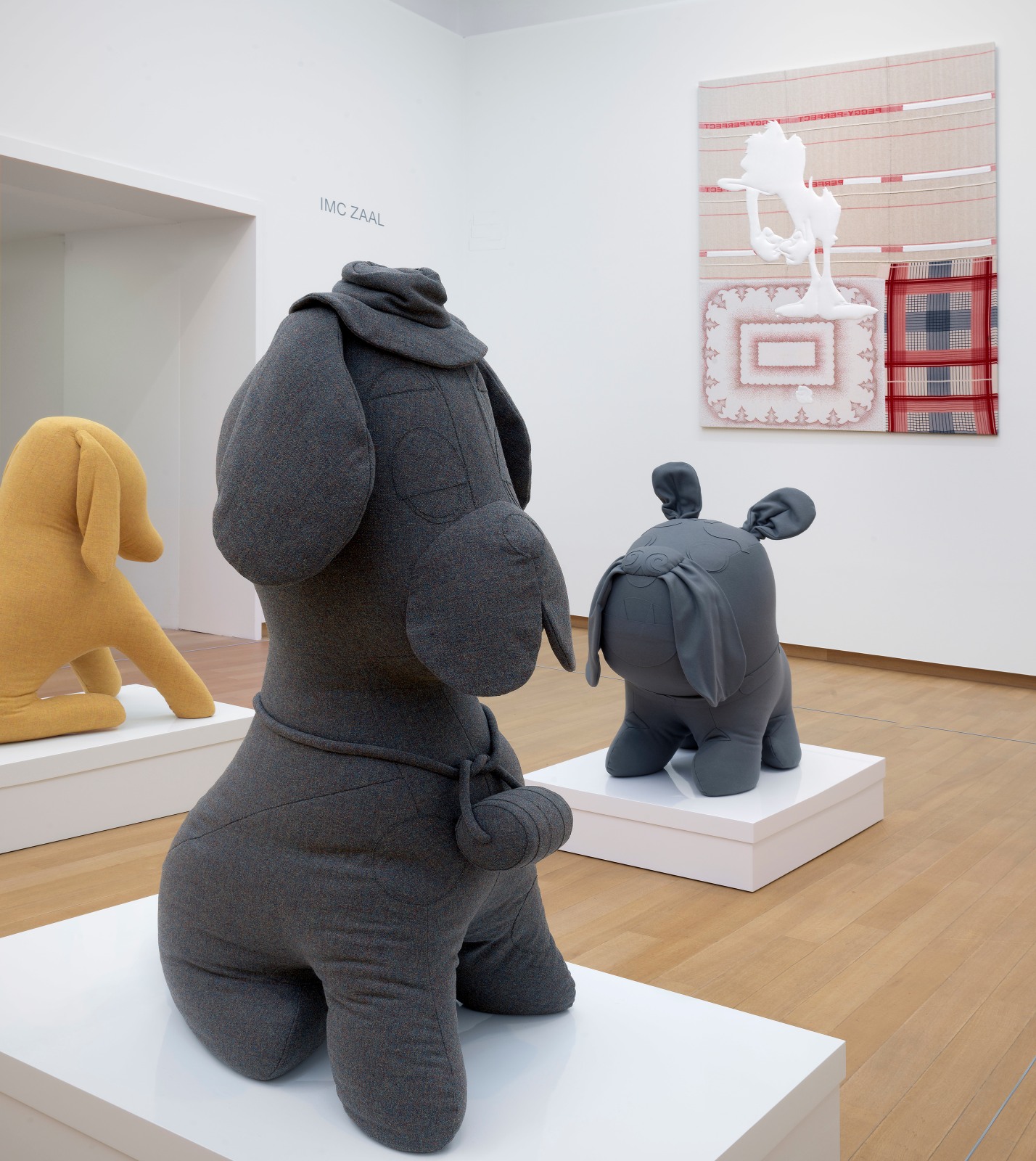 Installation view from the Stedelik Museum of three stuffed dogs. One is a gray Saint Bernard with a brimmed fabric hat on a white platform. Behind it is a stuffed gray bulldog on a platform and off to the side is a yellow stuffed flappy-eared dog. Hanging on the wall in the background is a fabric painting.