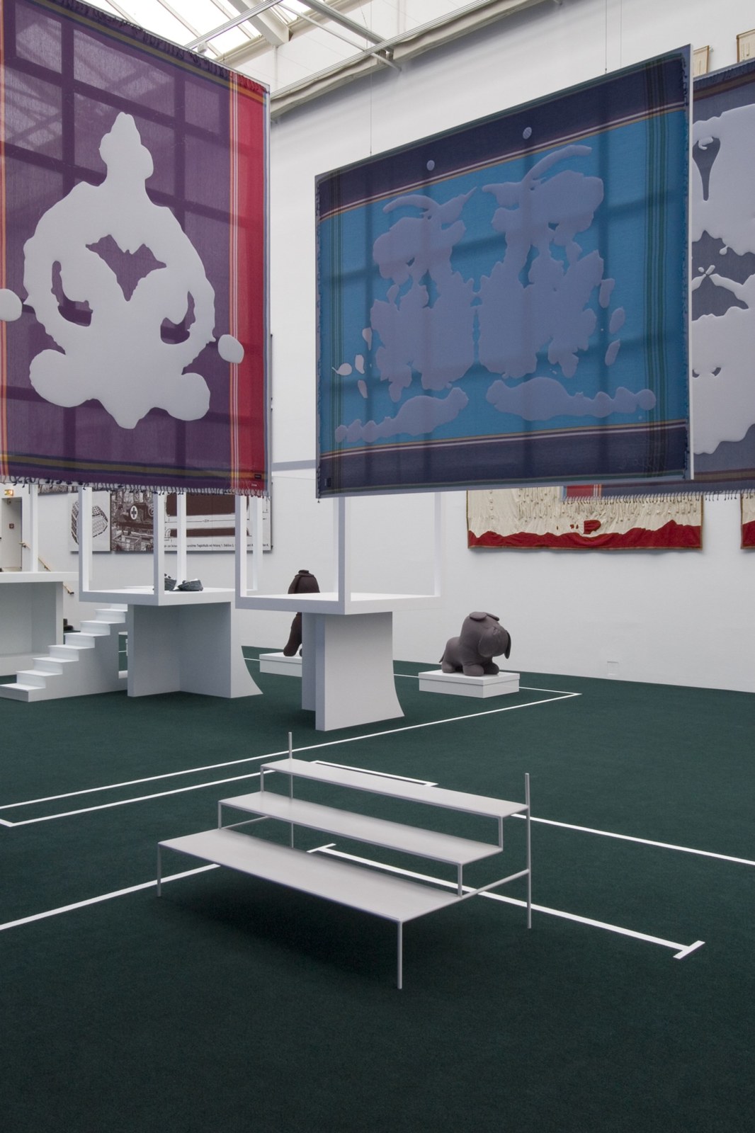Installation view from documenta 12 featuring two of von Bonin's rorschach test fabric paintings. One is pink and purple with the white rorschach blob appliqu&eacute;d on, and the second is light and dark blue with the white rorscah test appliqu&eacute;d on. The paintings are hanging from the ceiling, below them are small white bleachers and in the background are white cubes on platforms and a large gray stuffec bulldog on a white platform.