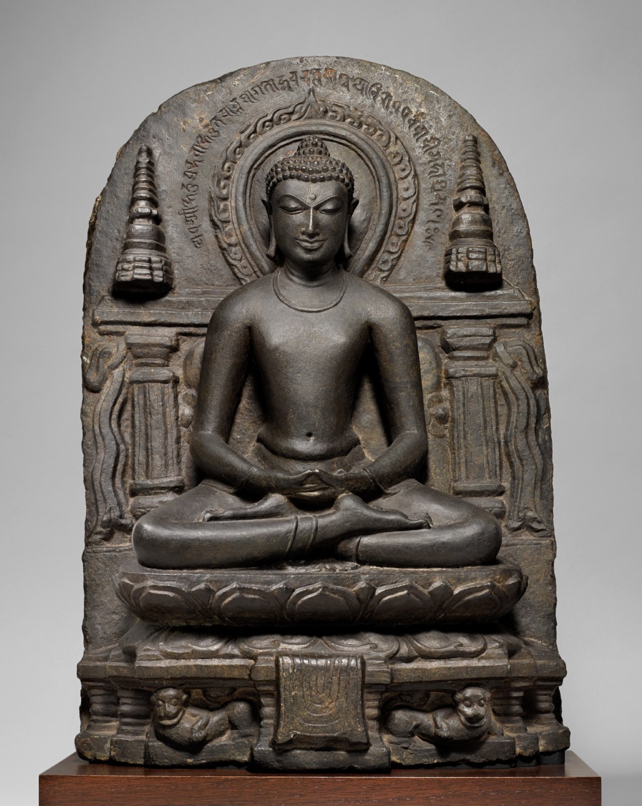 In this elegant and serene stele, the central figure of Buddha is flanked by two cylindrical stupas and is seated atop a lotus pedestal that in turn rests upon a lion throne.