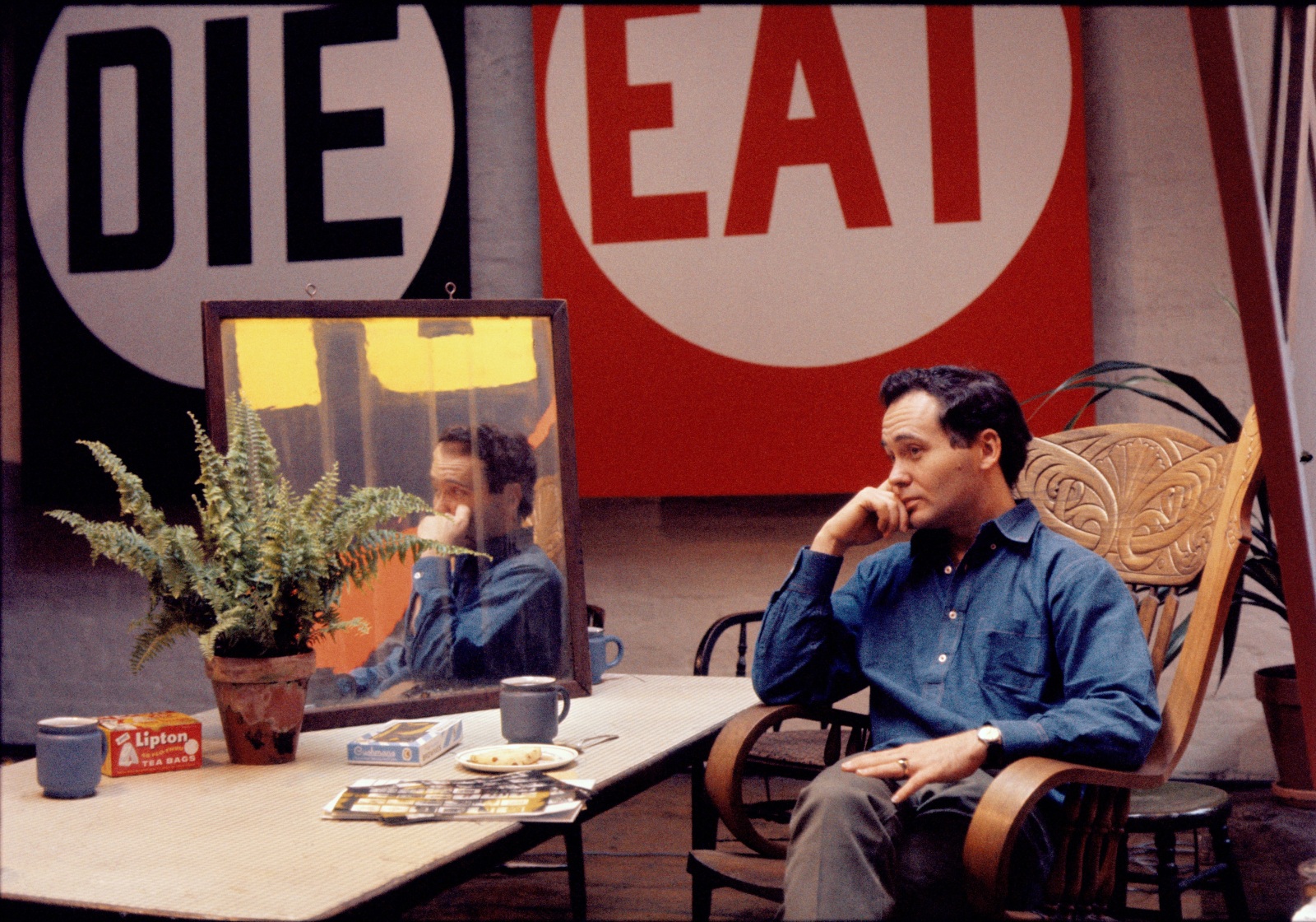 A photograph of Robert Indiana seated. wearing a blue shirt, by a table with a mirror reflecting his image and part of painting panel The Red Diamond Die. Behind him is the painting Eat/Die, a diptych with one canvas consisting of the red word eat in a white circle against a red ground, and the other the black word die in a white circle against a black ground.