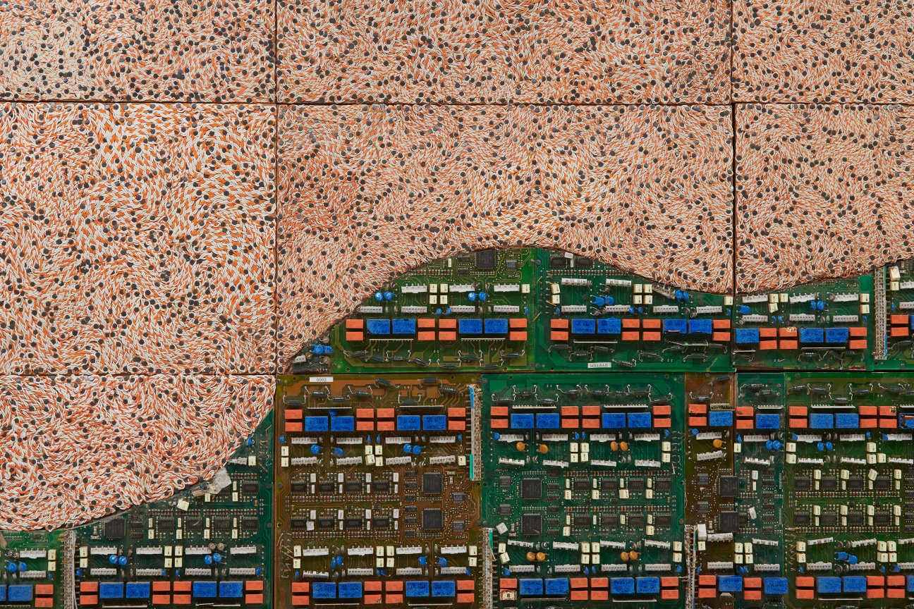 half of the image is composed of swirling orange electrical wires while the other is a mostly green surface of other types of electrical components