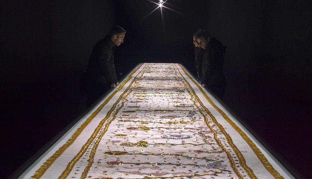 a dark room containing a light shining an embroidered canvas saturated with blood; the embroidery consists of golden fringes and beads