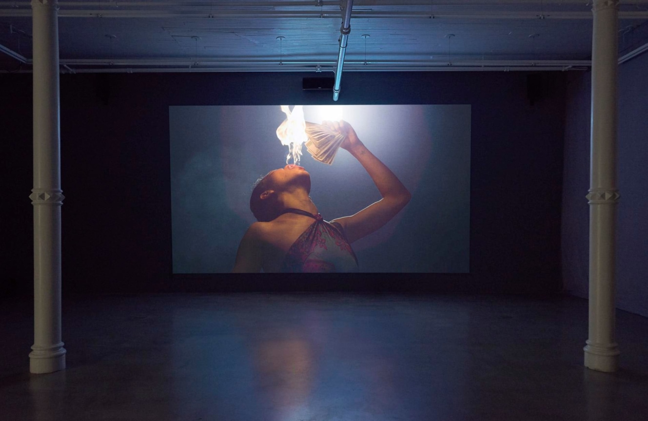 installation view of a woman breathing fire, setting money aflame