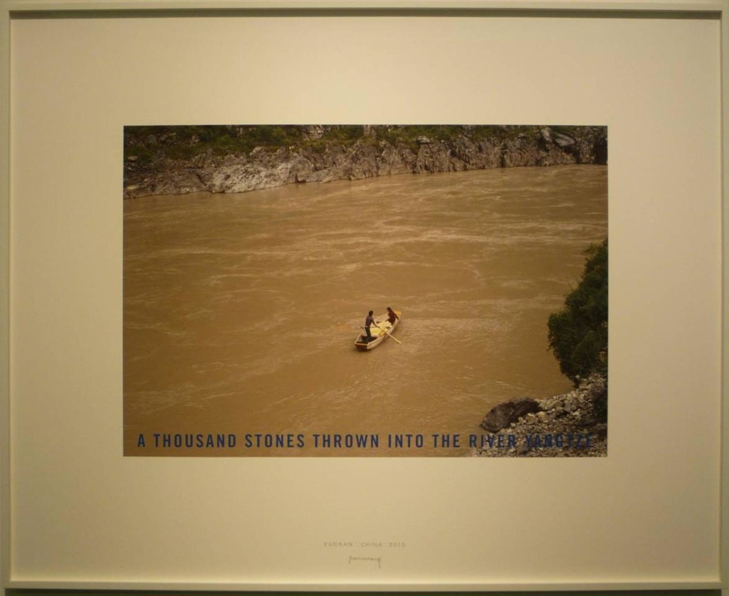 photograph of two figures riding a solitary rowboat in the middle of a brown river