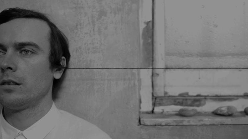 grayscale still of video of a man standing to the side of a dirty window