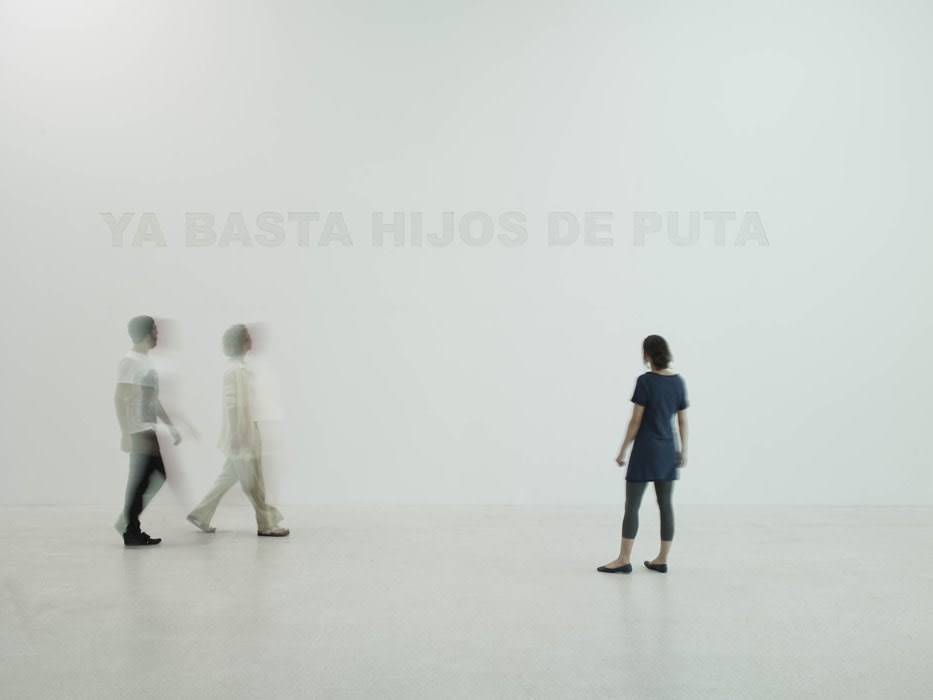 two people are walking by and another stops to look at the carved words saying &quot;YA BASTA HIJOS DE PUTA&quot; on a white wall