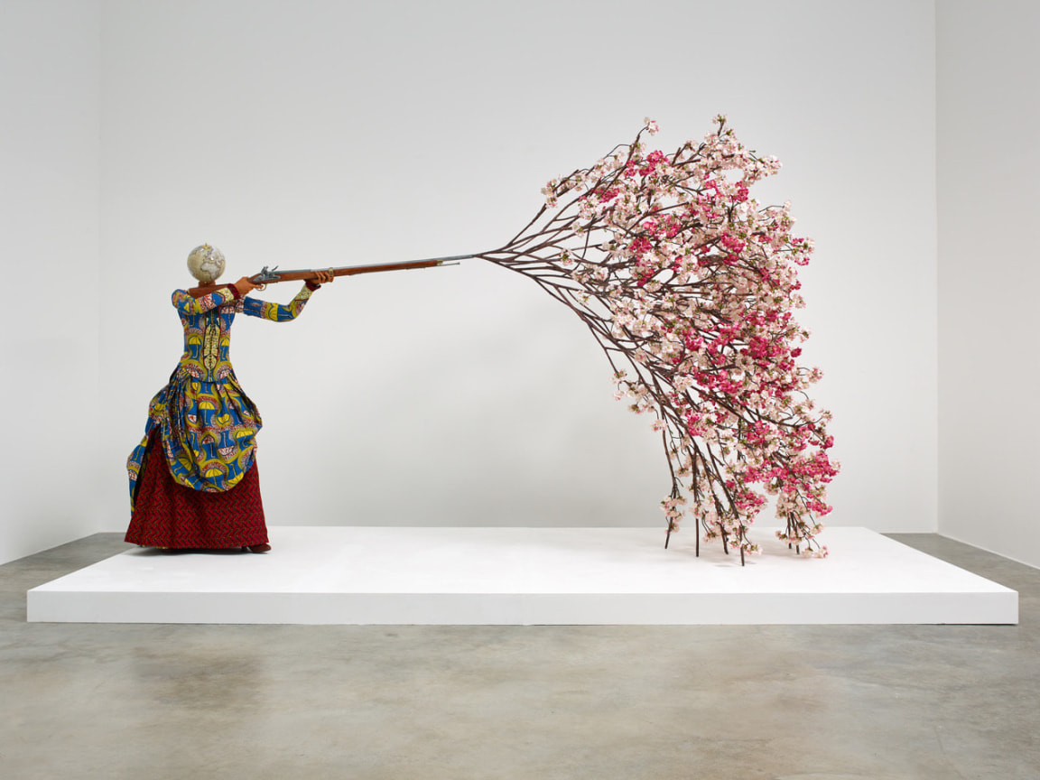 mannequin of a woman with a globe for a head holding a rifle that is shooting out a cherry blossom tree