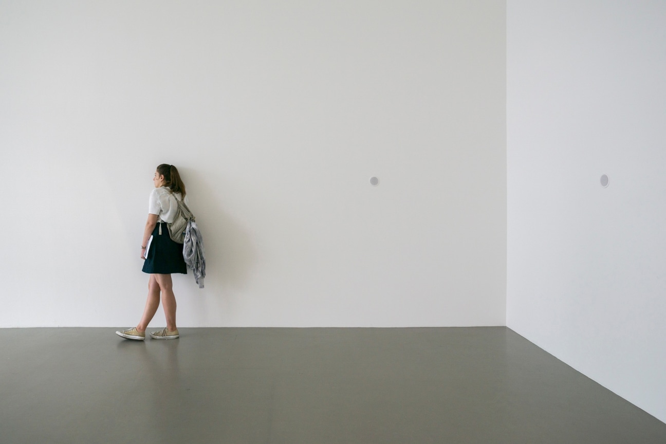 Woman leans against wall to listen to audio piece from a speaker which tells the story of the person who created the embroidery work displayed nearby