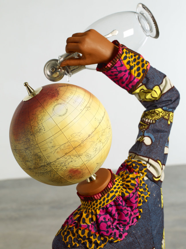 close up of a mannequin with a globe for a head pouring a drop of water from a pitcher on herself