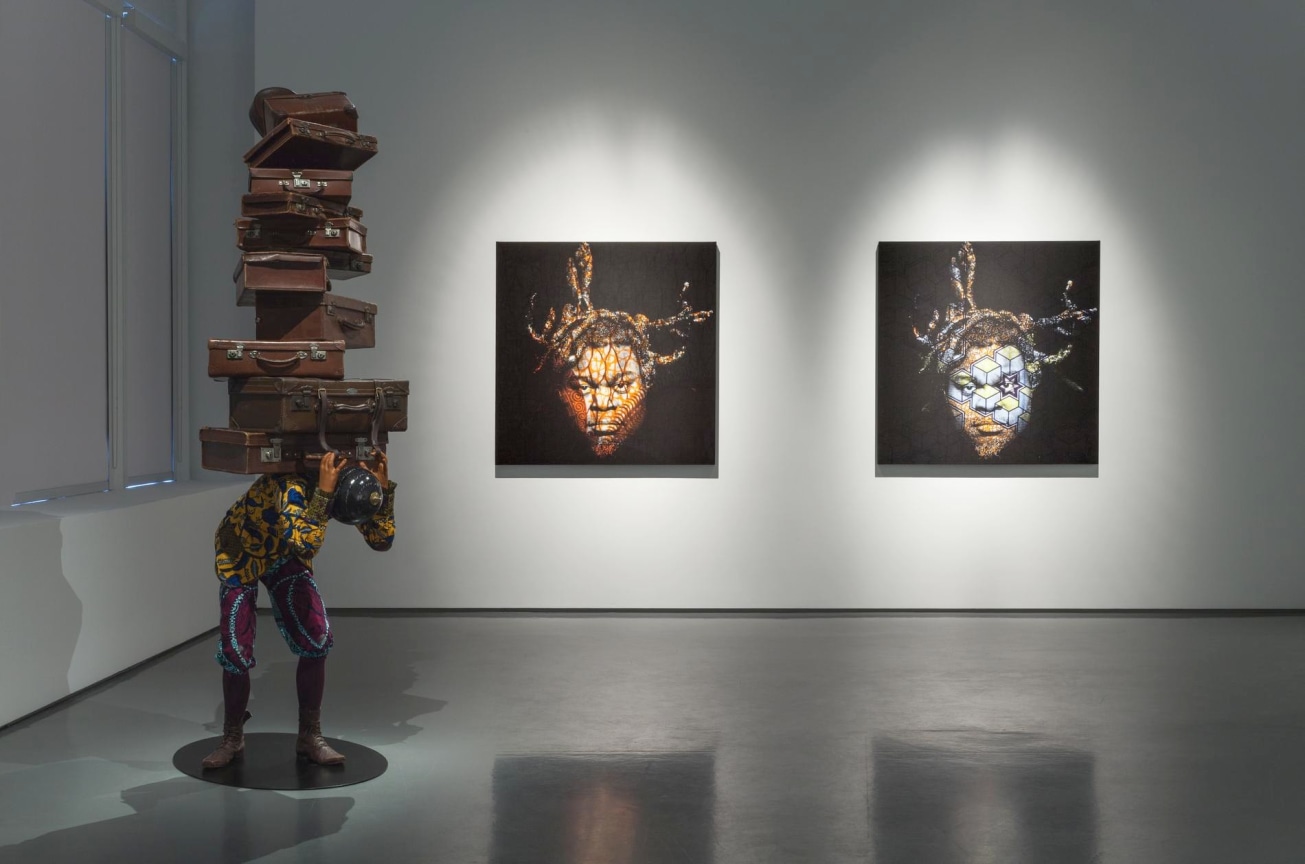 installation view of a mannequin carrying many suitcases on his back alongside two portraits for the same man