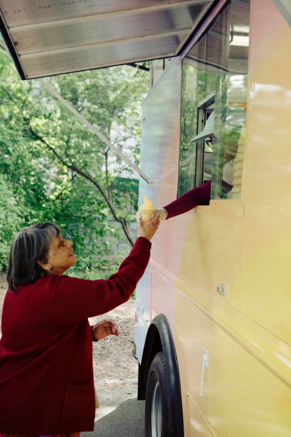 elderly woman being handed over yellow ice cream on a waffle cone by an employee at the solar powered, pastel colored, ice cream truck