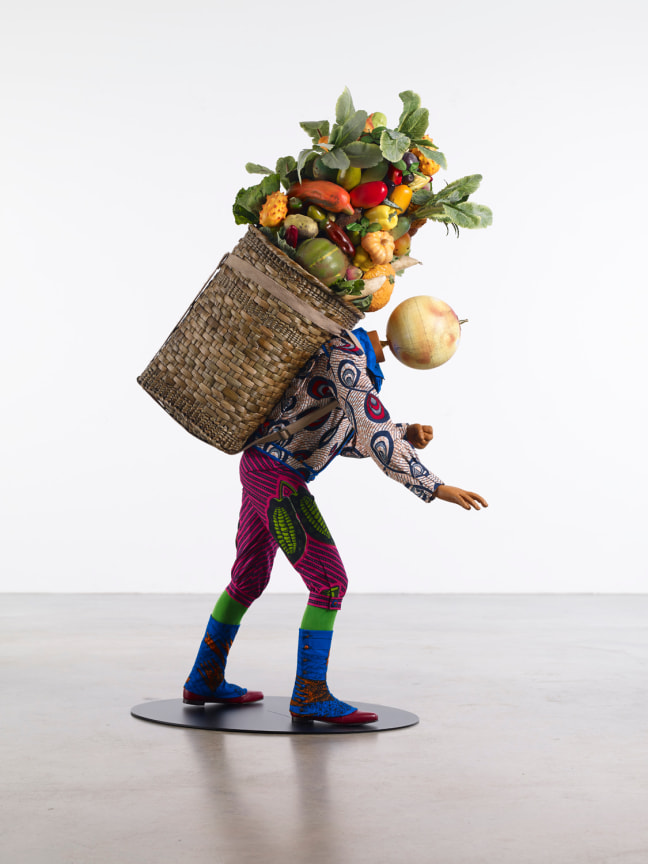 a mannequin of a child with a globe for a head carrying a mound of vegetables on a reed basket on his back