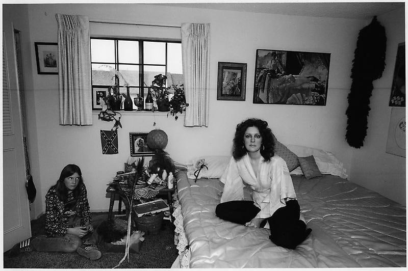 beautiful woman with voluminous hair sitting on he bed with her daughter giving her a aide eye from the floor