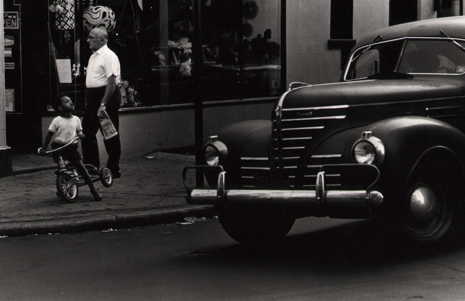 16. Anthony Barboza, NYC, 1970s. A dark vehicle fills the right of the frame. A man in a white shirt walks on the sidewalk while a young boy rides a tricycle in front of him.